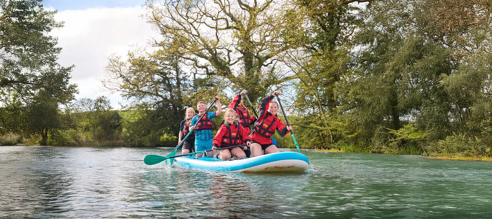 Four people wearing life jackets paddle on an inflatable board on a river, surrounded by trees.