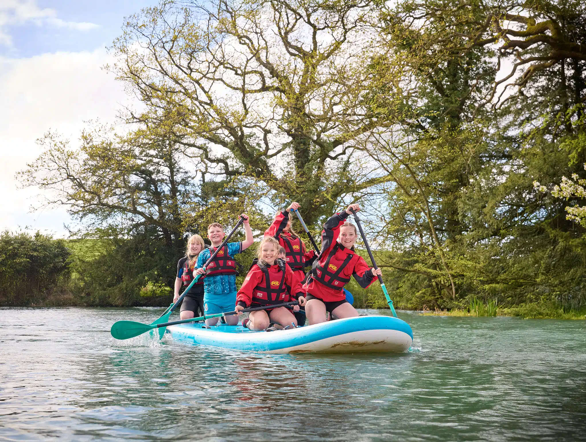 Four people in life jackets paddle a large inflatable board on a calm river, surrounded by trees.