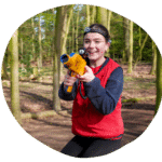 Person outdoors in a wooded area wearing a red vest and headgear, holding a toy laser tag gun, smiling.