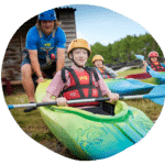 Person assisting a child in a kayak. Both are wearing helmets and personal flotation devices. Other people are visible in the background near water.