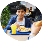 A boy sitting at a table eats a burger and fries from a blue tray in a cafeteria, with a yellow cup beside him. A plant is visible in the background.