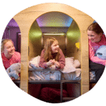 Three girls in colorful hoodies smile and chat while sitting on bunk beds in a cozy room with warm lighting.