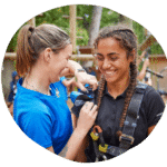 Two women interacting, one adjusting safety gear on the other. Both are smiling. Outdoor setting with climbing ropes in the background.