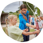 An instructor helps a young girl with a bow and arrow. Other children watch in the background during the outdoor archery session.