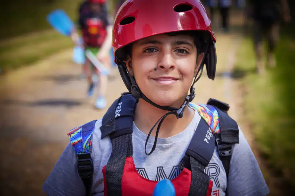 Person wearing a red helmet and life vest, holding a blue paddle. Outdoor setting with blurred figure in the background.