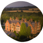 Aerial view of a large, old brick mansion surrounded by trees and open fields under a clear sky.