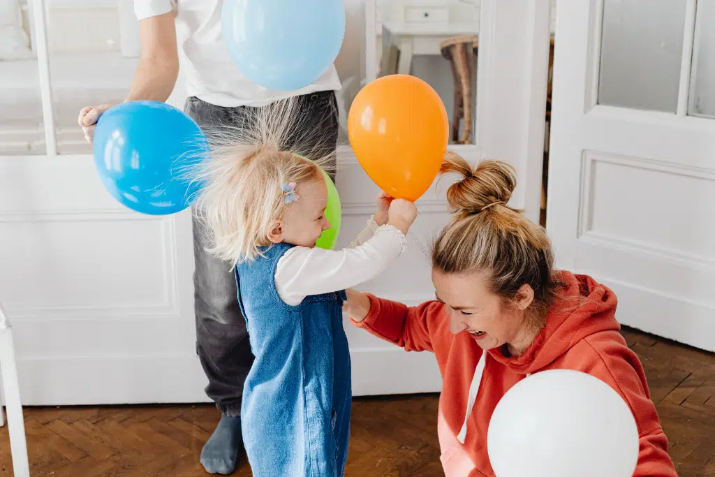 A toddler holding balloons with static hair stands next to a smiling woman kneeling on the floor; another person is partially visible holding a blue balloon.