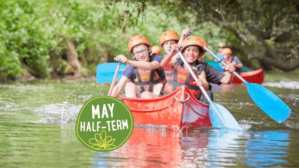 Four children wearing helmets and life jackets paddle a red canoe on a river, highlighting the excitement of PGL Adventure Holidays, with a 