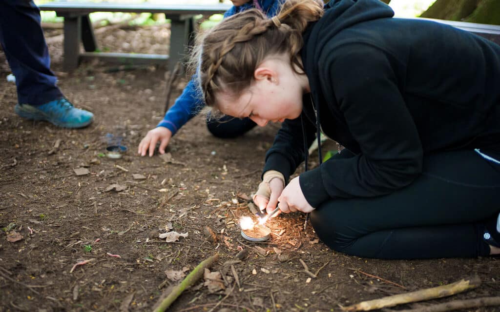 A girl crouches on the ground outdoors at PGL Adventure Holidays, using a flint and steel to start a small fire, while another person sits nearby.