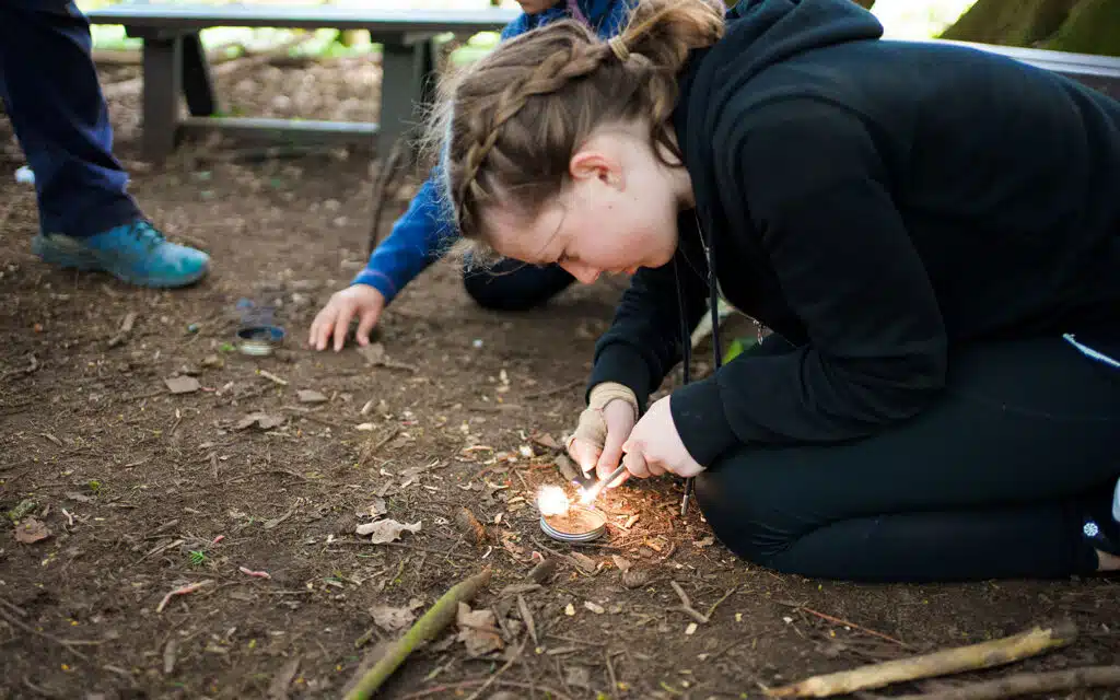 A girl crouches on the ground outdoors at PGL Adventure Holidays, using a flint and steel to start a small fire, while another person sits nearby.
