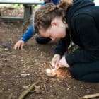 A girl crouches on the ground outdoors at PGL Adventure Holidays, using a flint and steel to start a small fire, while another person sits nearby.