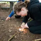 A girl crouches on the ground outdoors at PGL Adventure Holidays, using a flint and steel to start a small fire, while another person sits nearby.