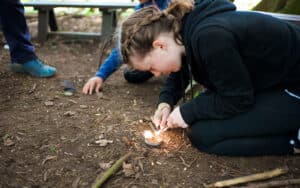 A girl crouches on the ground outdoors at PGL Adventure Holidays, using a flint and steel to start a small fire, while another person sits nearby.