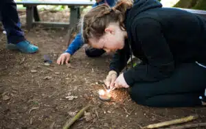 A girl crouches on the ground outdoors at PGL Adventure Holidays, using a flint and steel to start a small fire, while another person sits nearby.