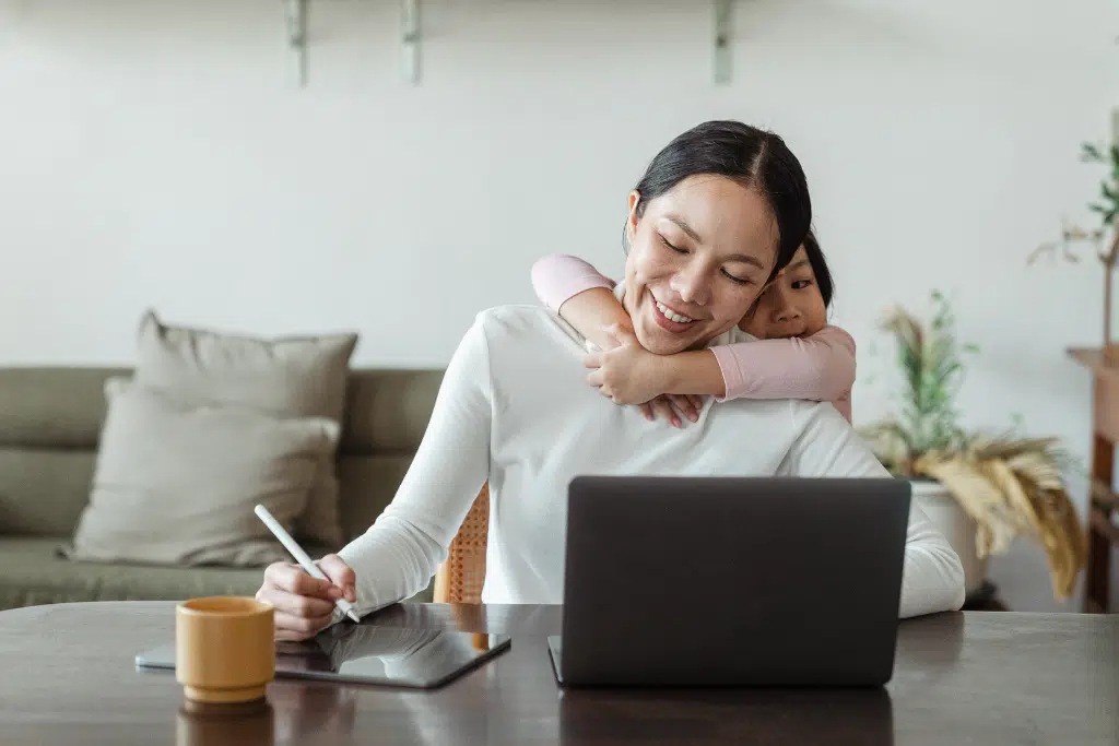 A woman works on a laptop and tablet at a table while a young child hugs her from behind in a home setting.