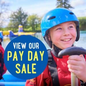 A child wearing a blue helmet and red life jacket smiles while holding a paddle near water. Text reads, “View our Pay Day Sale.”.