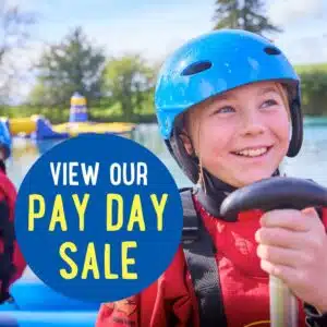 A child wearing a blue helmet and red life jacket smiles while holding a paddle near water. Text reads, “View our Pay Day Sale.”.