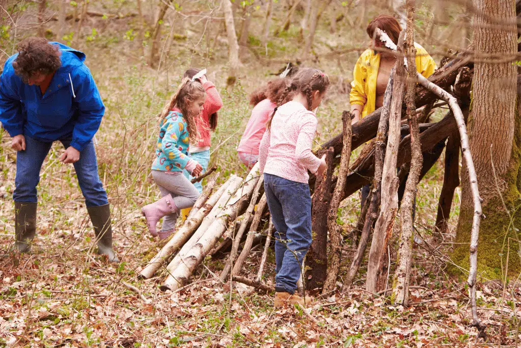 A group of children and two adults build a structure with sticks and logs in a wooded area.