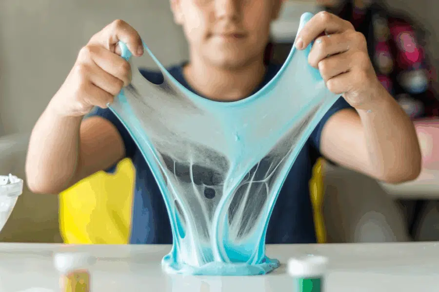 A child stretches blue slime with both hands over a white table, with small containers of crafting supplies in the foreground.