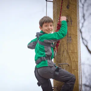 A boy in a harness climbs a wooden outdoor pole, holding onto a red rope, with a smile on his face.
