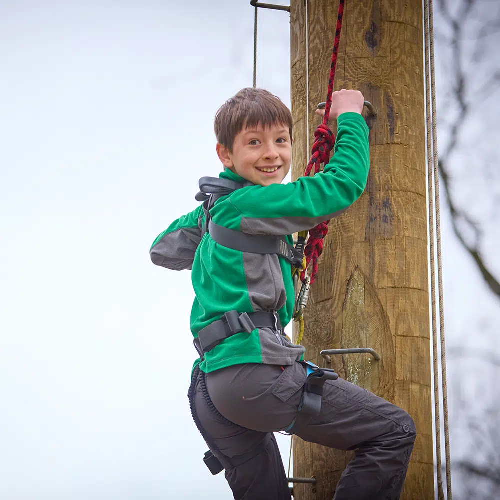 A boy in a harness climbs a wooden outdoor pole, holding onto a red rope, with a smile on his face.