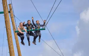 Three people wearing harnesses sit on a suspended swing, holding on to ropes with their legs dangling, against a blue sky background.