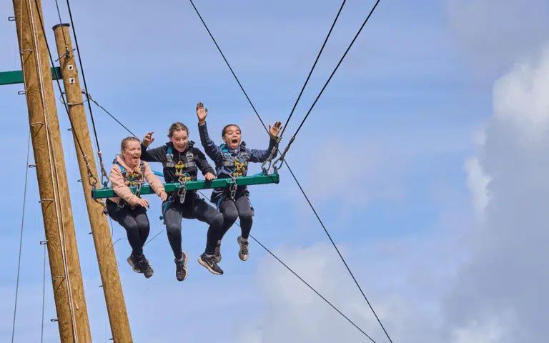 Three people wearing harnesses sit on a suspended swing, holding on to ropes with their legs dangling, against a blue sky background.