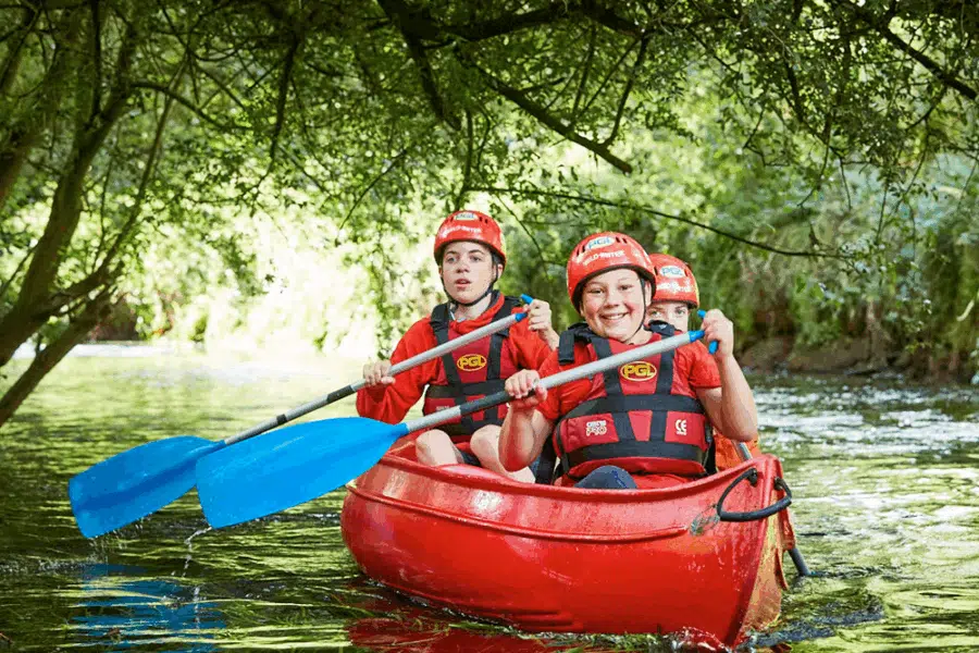Three children wearing helmets and life jackets paddle a red canoe with blue oars under tree branches on a calm river.