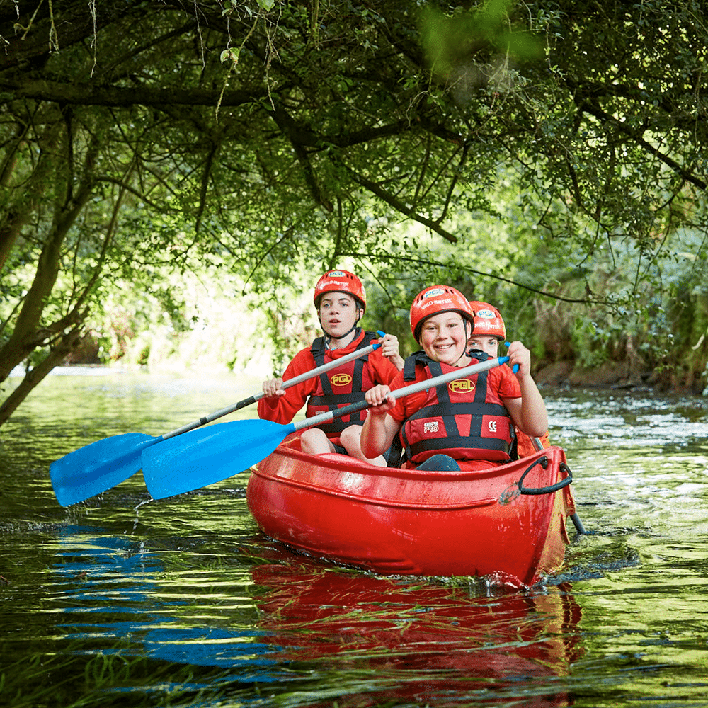 Three children wearing helmets and life jackets paddle a red canoe with blue oars under tree branches on a calm river.