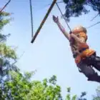 A child in a harness reaches for a wooden bar during the Pony Camp Improver at Liddington on July 27, 2024, with trees and blue sky in the background.