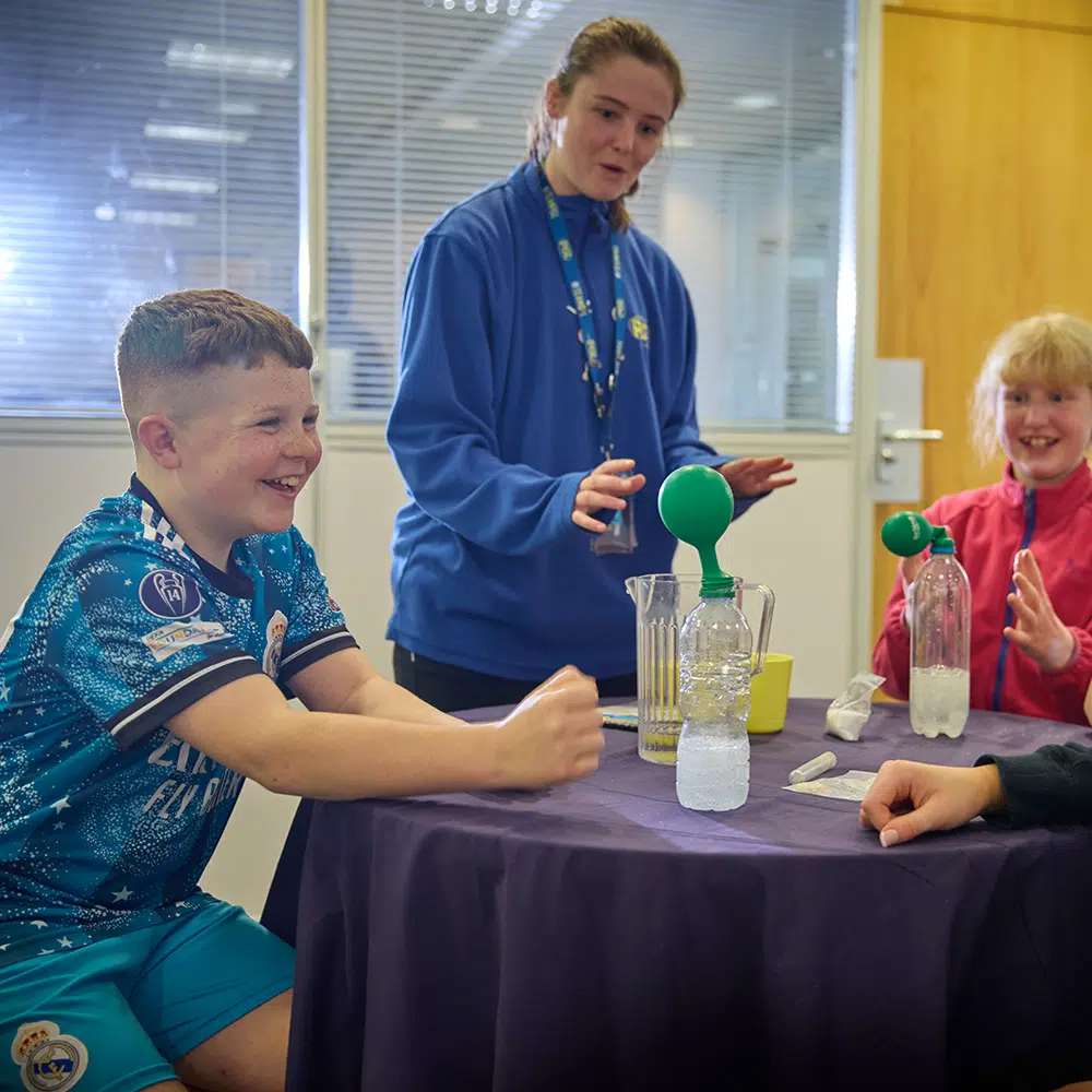 Three children sit around a table doing a science experiment with bottles, balloons, and water as one adult stands nearby, all appearing engaged and observant.