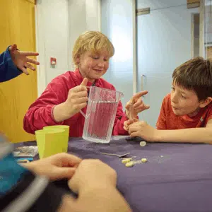 Two children sit at a table with a clear pitcher, cups, and small objects, appearing to engage in a hands-on activity or experiment.