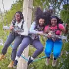 Three young women enjoy the Learner Driver event at Liddington on Aug 10, 2024, smiling and laughing as they're suspended in harnesses on an outdoor ride with trees in the background.