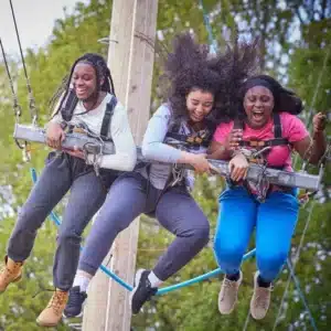 Three young women enjoy the Learner Driver event at Liddington on Aug 10, 2024, smiling and laughing as they're suspended in harnesses on an outdoor ride with trees in the background.