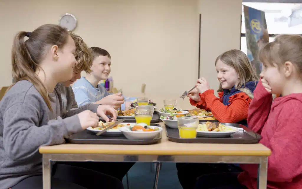 Five children sit at a table in a cafeteria, eating meals from trays and talking together.