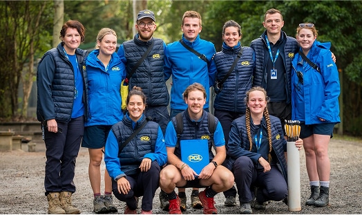 A group of ten people wearing matching blue and navy PGL uniforms pose outdoors on a gravel path, surrounded by trees.