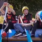 Three children in helmets and life jackets smile and paddle on an inflatable raft during a Cool Science activity at Liddington, July 20, 2024.