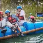 Four people in helmets and life vests sit on a blue raft on the water at Aqua Explorer, Bawdsey Manor (Jul 20, 2024), surrounded by trees, with another person visible in the background.