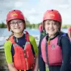 Two children in red helmets, life jackets, and wetsuits smile by the water at Aqua Explorer, Bawdsey Manor – Jul 20, 2024. Boats and people can be seen in the background.
