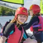Two young people in red helmets, life jackets, and wetsuits sit on a small sailboat at Aqua Explorer, Bawdsey Manor (Jul 20, 2024); one smiles at the camera while the other looks ahead.