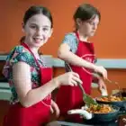 At Cooks Academy, Liddington (Aug 03, 2024), two girls in red aprons cook food on a stovetop; one smiles at the camera while the other concentrates on her cooking.