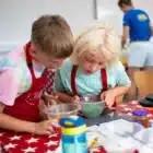 At Cooks Academy, Liddington on August 3, 2024, two children in aprons examine a bowl of green peas on a red star-pattern tablecloth while another person stands in the background.