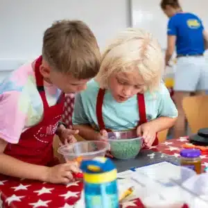 At Cooks Academy, Liddington on August 3, 2024, two children in aprons examine a bowl of green peas on a red star-pattern tablecloth while another person stands in the background.
