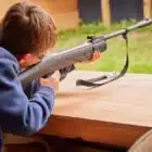 At Boreatton Park’s Pony Camp Beginner on July 20, 2024, a person with short brown hair aims a rifle while seated at a wooden table at an outdoor shooting range.