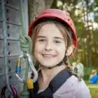 A young girl in climbing gear smiles outdoors next to ropes and carabiners at Bake It, Liddington – Jul 27, 2024.