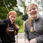 Two smiling girls with red hair in braids, wearing casual clothes, stand outdoors among trees, one giving a thumbs up at Cooks Academy, Liddington - Aug 03, 2024.