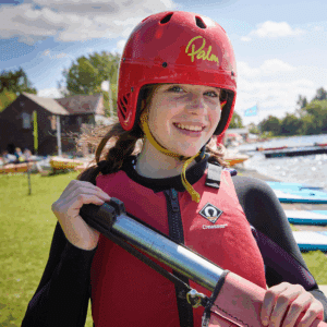 A young person wearing a red helmet and life jacket holds paddle gear by a lakeside with kayaks and canoes in the background.