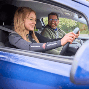 A young woman in the driver’s seat smiles while holding the steering wheel, with an older man smiling beside her in the passenger seat of a blue car.