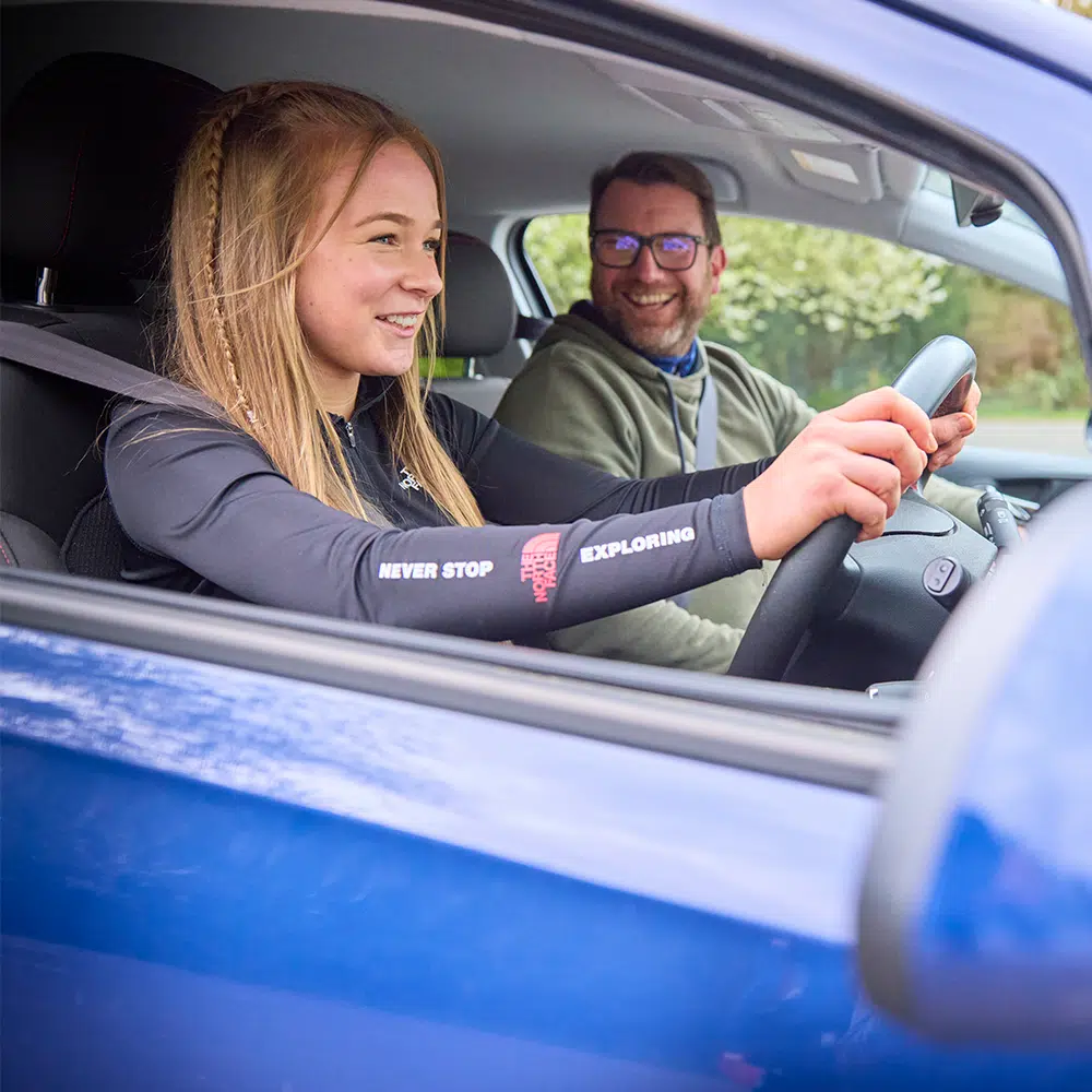 A young woman in the driver’s seat smiles while holding the steering wheel, with an older man smiling beside her in the passenger seat of a blue car.