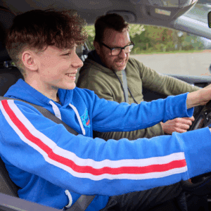 A teenage boy in a blue hoodie sits in the driver's seat of a car, smiling, while an adult man in glasses sits beside him, both wearing seatbelts.